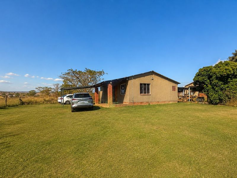 A detached single-story house featuring a front carport, brick pillar accents, and security bars on windows, set on a large open grass plot.