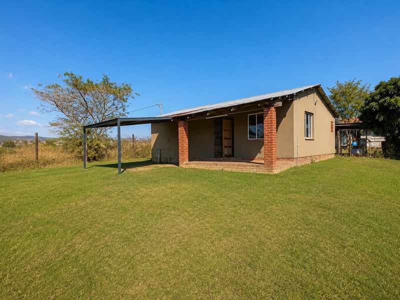 A detached single-story dwelling featuring a covered patio with brick pillars, a metal carport extension, and a large surrounding lawn area.