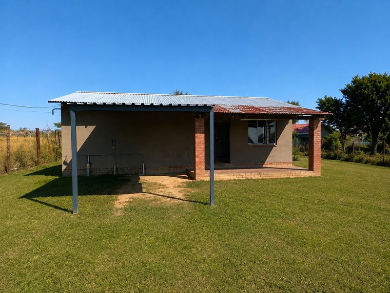 Rear view of a single-story house featuring a covered patio area, brick pillars, and a corrugated metal roof with visible signs of weathering.
