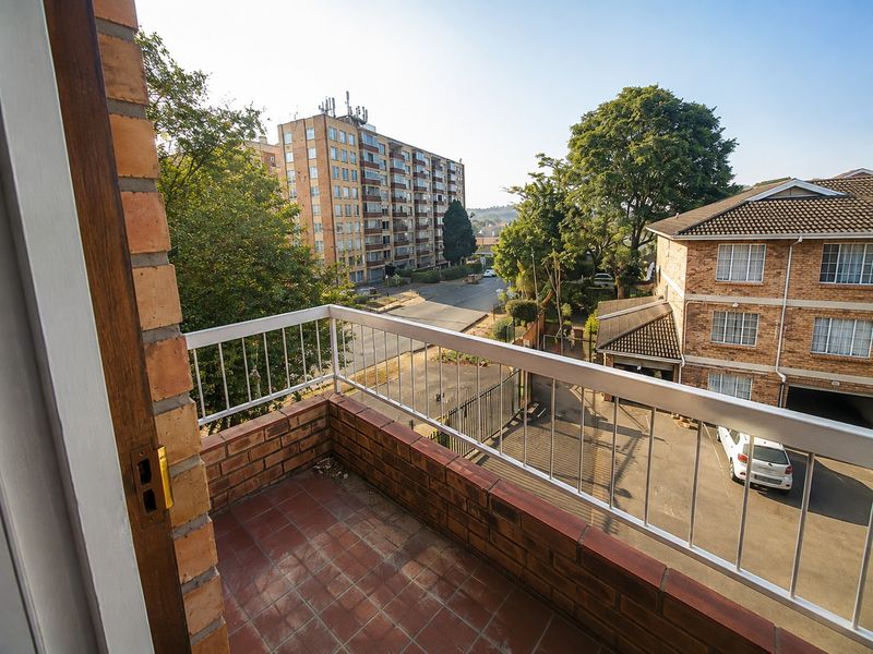 A private balcony featuring a brick balustrade with metal railing and tiled flooring, offering an elevated view of the surrounding apartment complex and street.