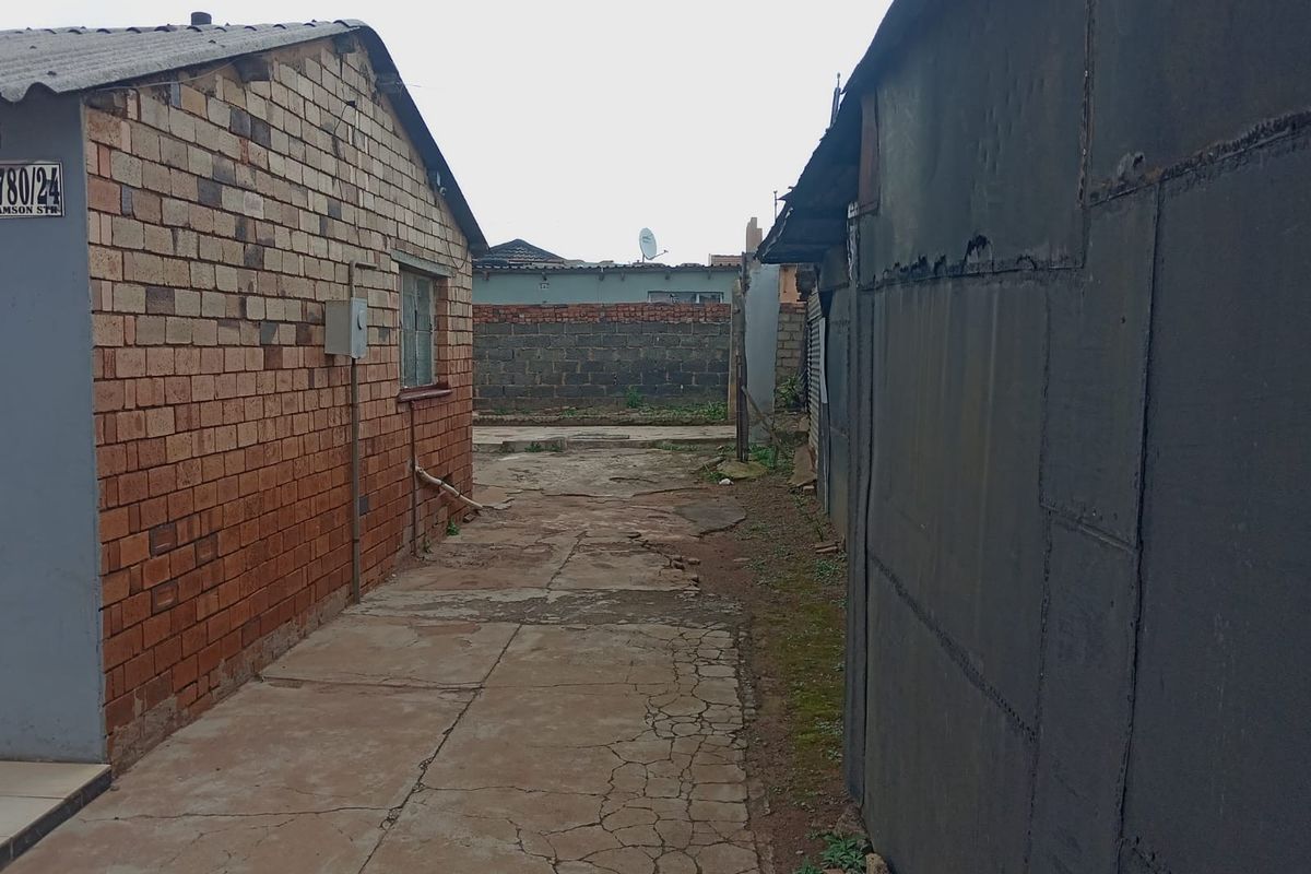 A narrow exterior side passage between two structures featuring weathered brick and metal wall finishes with cracked concrete paving.