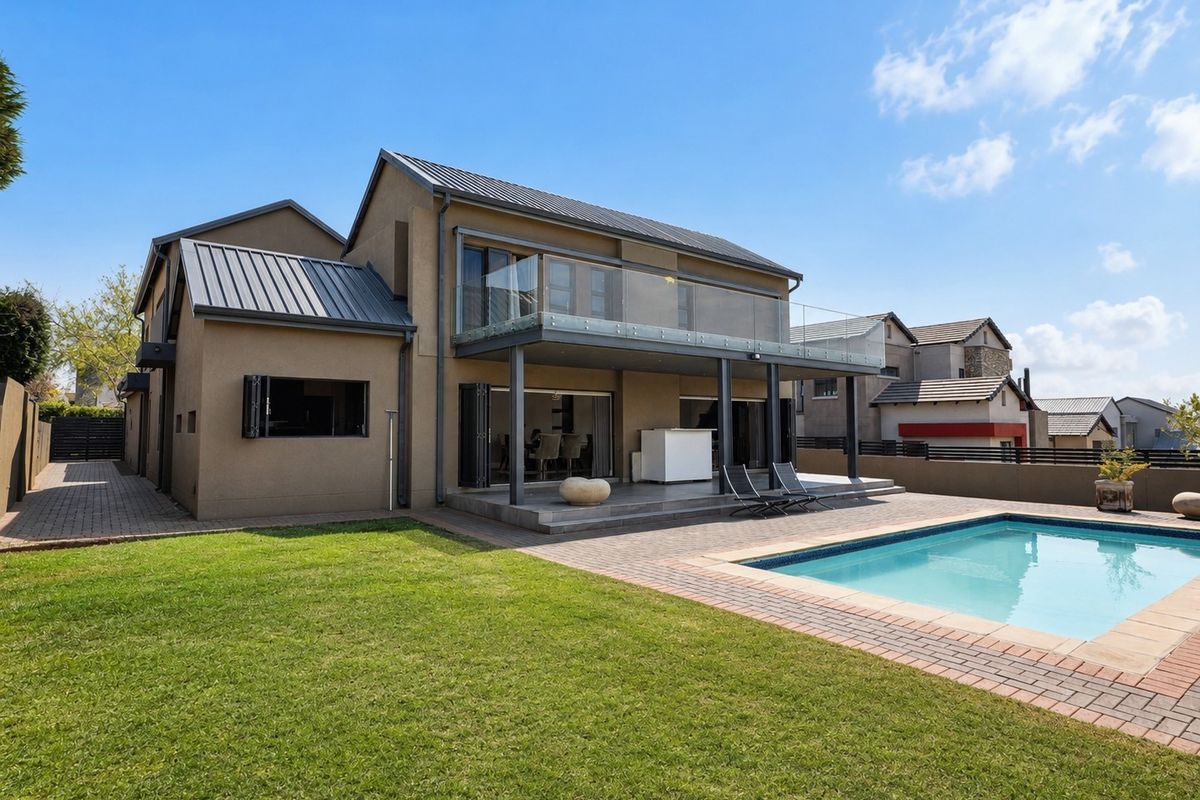 Rear view of a modern double-story home featuring a covered patio, glass-railed balcony, and a well-maintained swimming pool with paved surround.