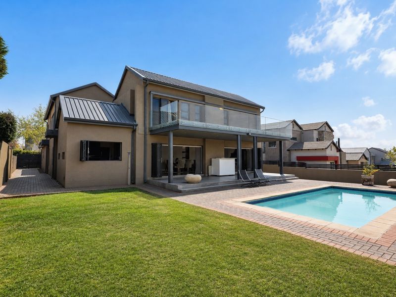 Rear view of a modern double-story home featuring a covered patio, glass-railed balcony, and a well-maintained swimming pool with paved surround.