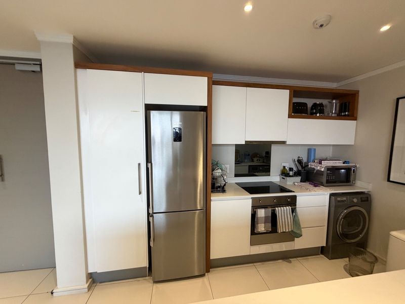 Compact modern kitchen featuring white cabinetry with wood-tone accents, integrated appliances, electric hob, built-in oven, and tiled flooring.