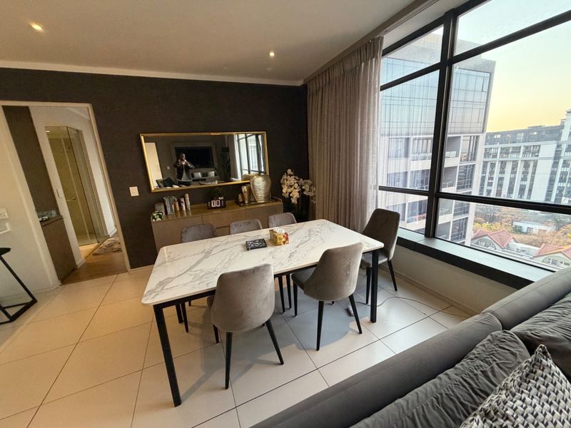 Modern dining area featuring large floor-to-ceiling windows with city views, neutral tiled flooring, and recessed lighting in a well-maintained apartment setting.