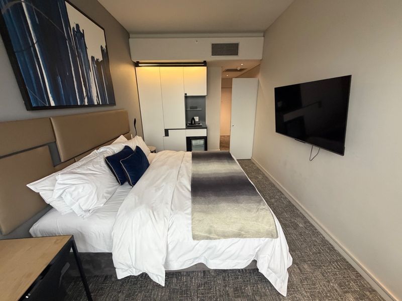 A modern bedroom featuring built-in cabinetry, a wall-mounted television, and an integrated air conditioning unit, finished with neutral tones and carpeted flooring.