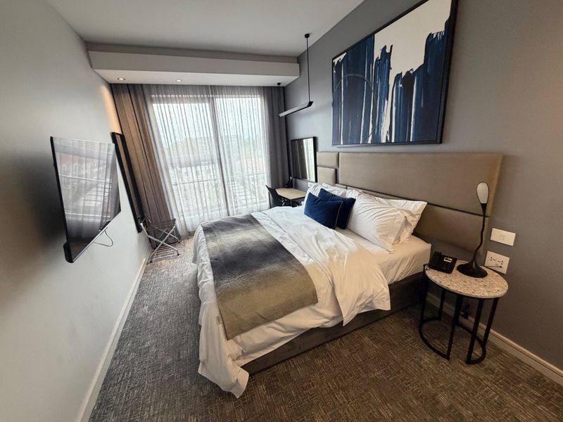A modern, well-appointed bedroom featuring a large upholstered headboard, recessed ceiling lighting, and a sliding glass door leading to a private balcony.