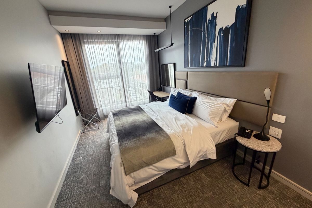 A modern, well-appointed bedroom featuring a large upholstered headboard, recessed ceiling lighting, and a sliding glass door leading to a private balcony.