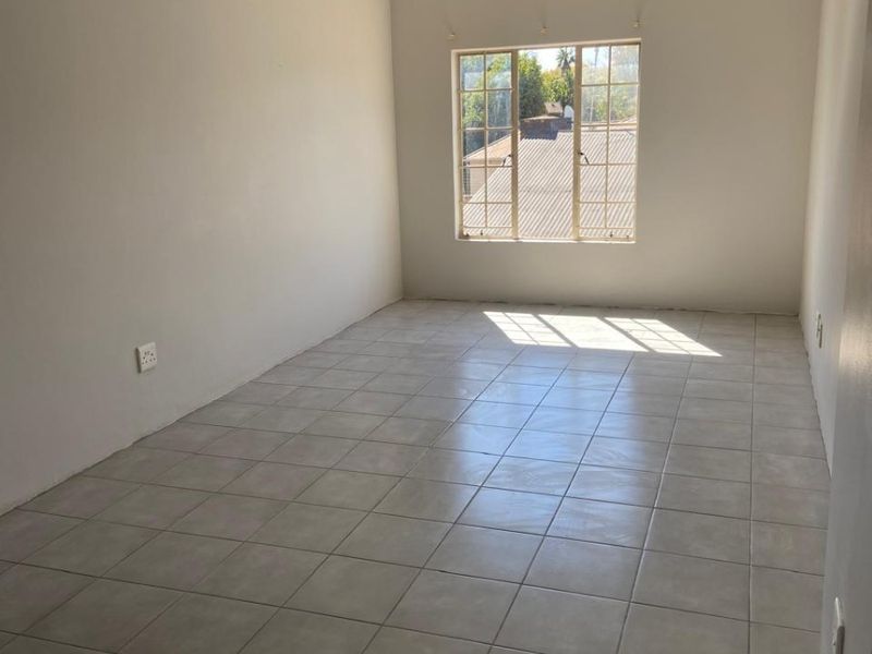 A bright, empty bedroom featuring neutral tiled flooring, a large window with security bars, and a simple ceiling-mounted spotlight fixture.