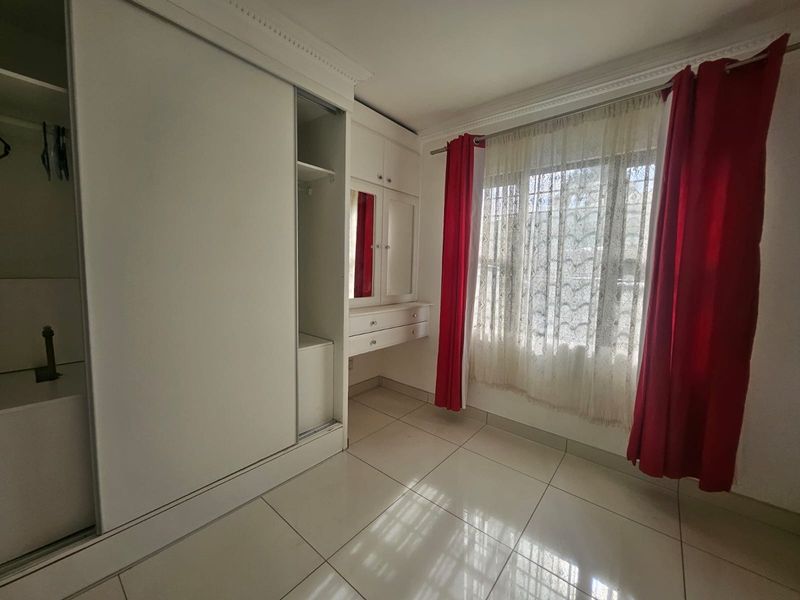 A bright bedroom featuring tiled flooring, built-in cupboards with an integrated vanity unit, and large windows with decorative curtains.
