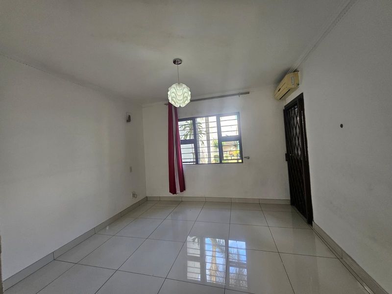 A bright, empty living room featuring tiled flooring, a wall-mounted air conditioning unit, and security bars on the window.