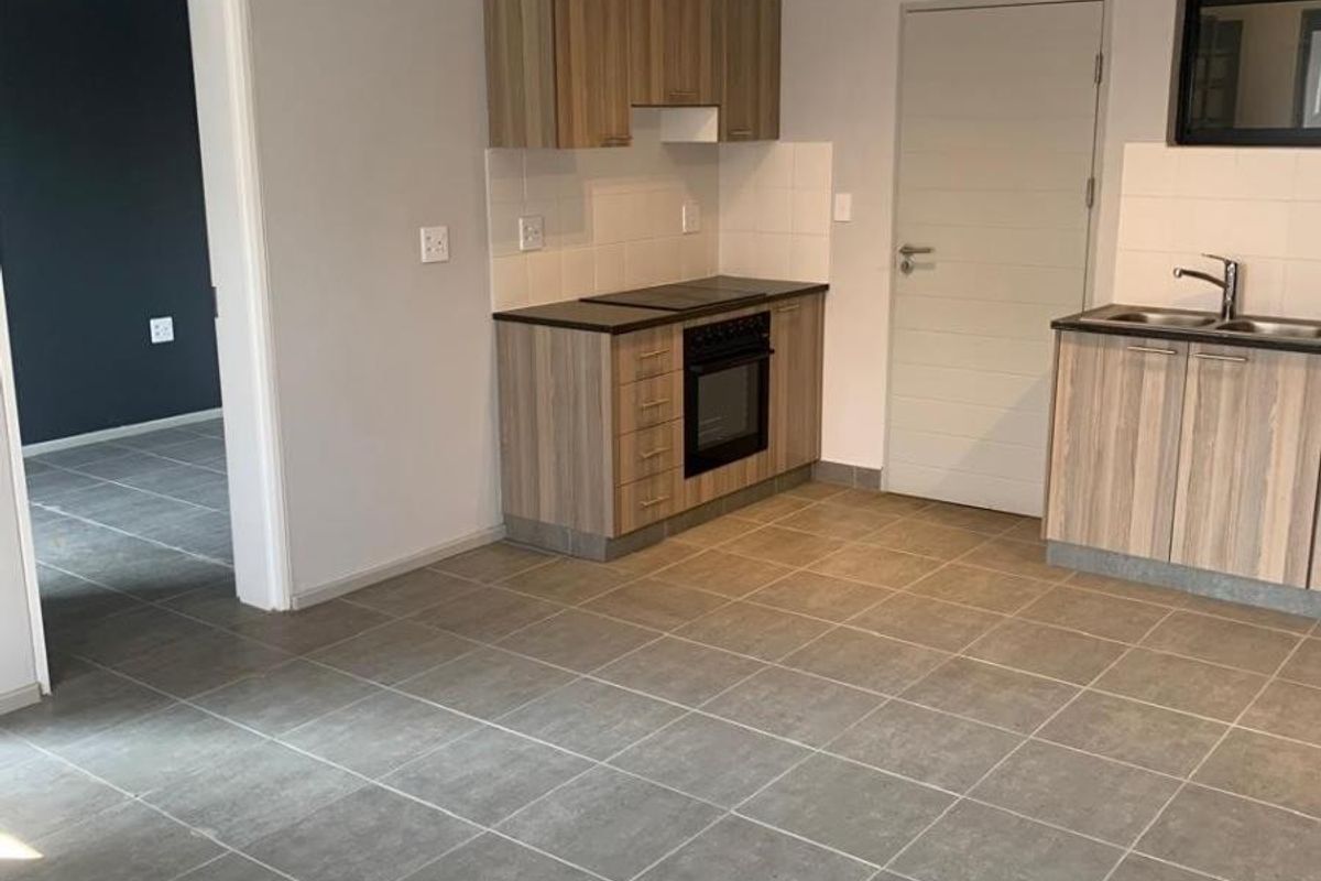 A clean, modern kitchen featuring light wood-tone cabinetry, built-in oven and hob, stainless steel sink, and neutral grey floor tiles.