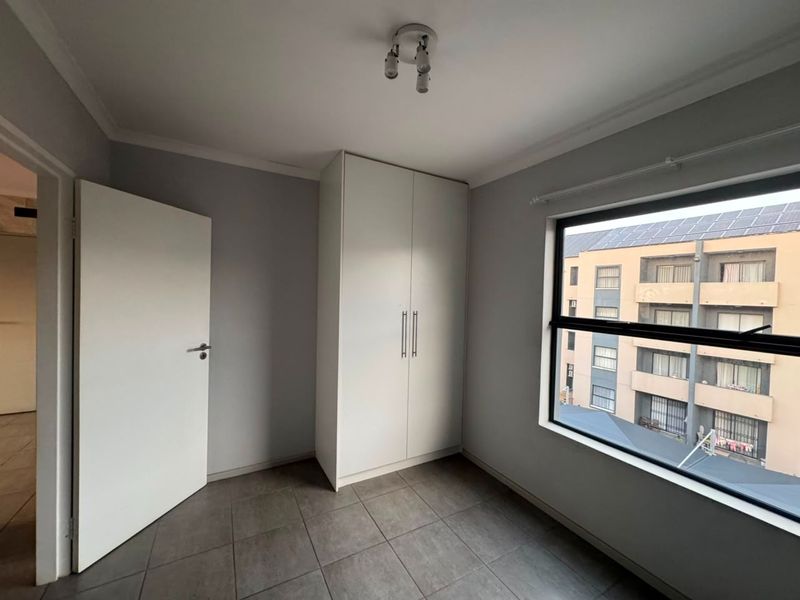 A clean, empty bedroom featuring neutral grey walls, tiled flooring, and a built-in white wardrobe with a view of the surrounding apartment complex.