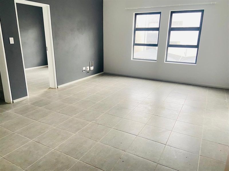A bright, empty living area featuring neutral tiled flooring, a dark grey accent wall, and large aluminium-framed windows allowing for natural light.