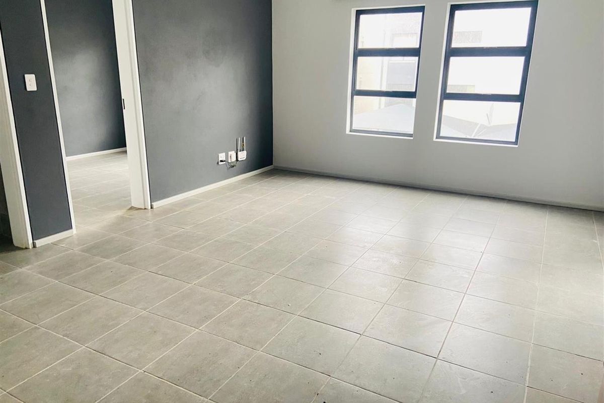 A bright, empty living area featuring neutral tiled flooring, a dark grey accent wall, and large aluminium-framed windows allowing for natural light.