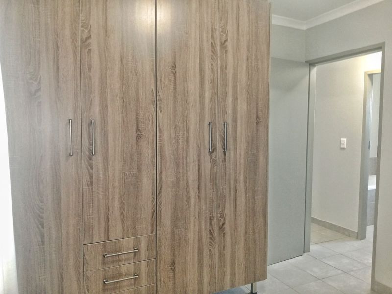 A bedroom featuring modern wood-grain built-in cupboards and neutral tiled flooring, with an adjacent doorway leading to an adjoining room.