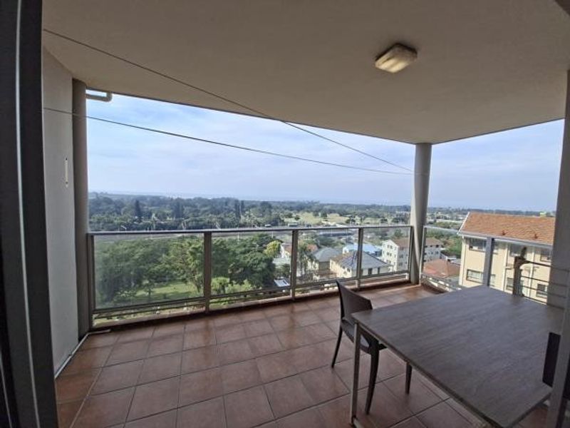 A spacious covered balcony featuring tiled flooring and a glass balustrade, offering expansive elevated views of the surrounding neighborhood and landscape.
