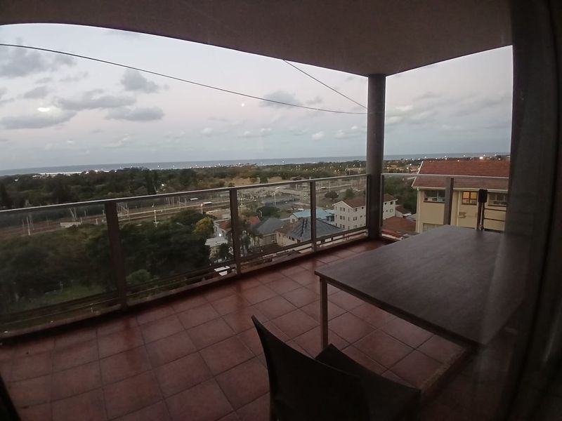 A covered balcony featuring tiled flooring and a glass balustrade, offering expansive elevated views of the surrounding neighborhood and coastline.