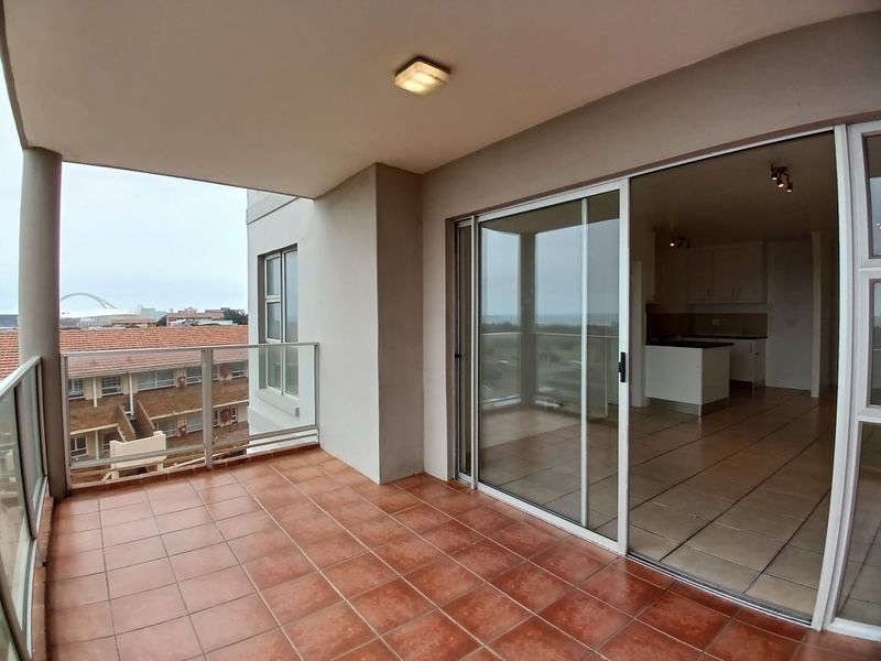 A spacious covered balcony featuring tiled flooring and a glass balustrade, offering seamless indoor-outdoor flow through large sliding doors into the open-plan living area.