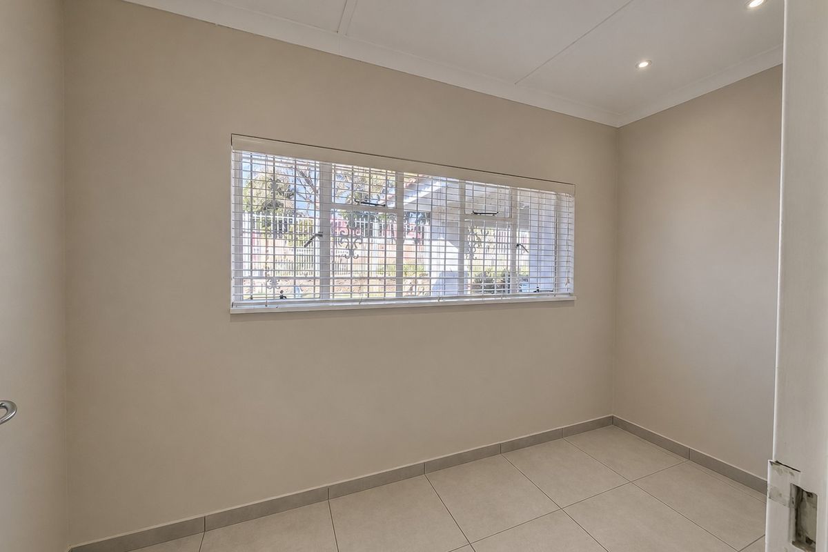 A clean, empty bedroom featuring neutral tiled flooring, recessed downlighting, and a large window fitted with security bars and venetian blinds.