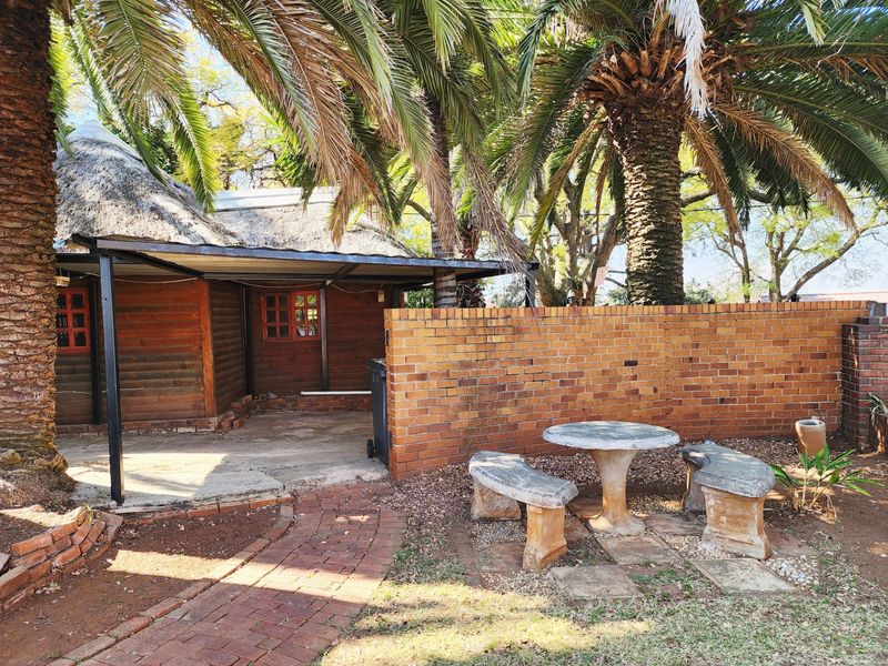 A rear exterior view featuring a thatched roof structure with timber cladding, a covered patio area, and a brick boundary wall in a garden setting.