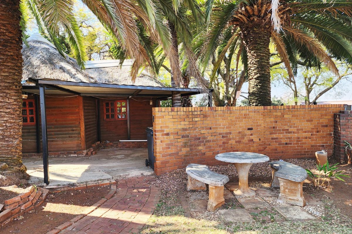 A rear exterior view featuring a thatched roof structure with timber cladding, a covered patio area, and a brick boundary wall in a garden setting.