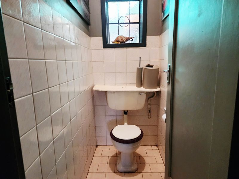 A compact, dated bathroom featuring ceramic wall and floor tiles, a standard toilet, and a small leaded glass window.