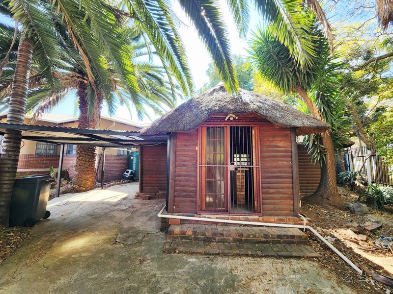 A detached timber log cabin with a thatched roof, featuring a security gate and adjacent metal carport on a concrete paved area.
