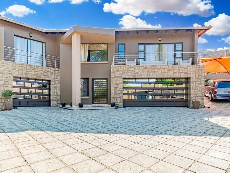Modern double-story residential facade featuring stone cladding, a spacious paved driveway, integrated double garages, and upper-level balconies with metal railings.