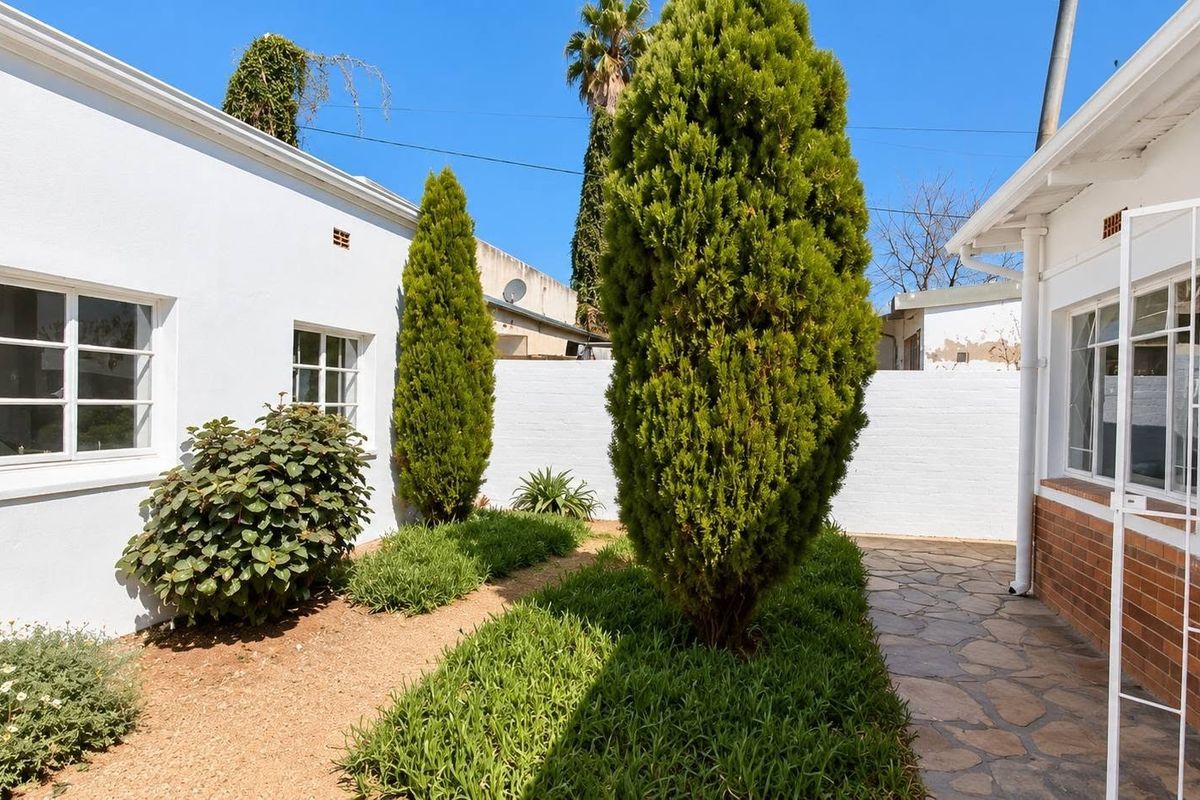 A well-maintained rear courtyard featuring stone paving, landscaped garden beds with mature shrubs, and white masonry walls connecting the building sections.