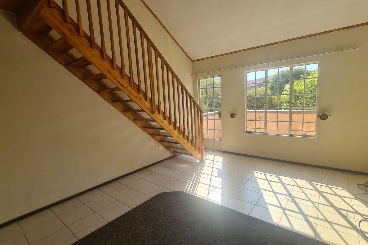 An open-plan living area featuring a wooden staircase, tiled flooring, and natural light through large windows and a glass door leading to a balcony.