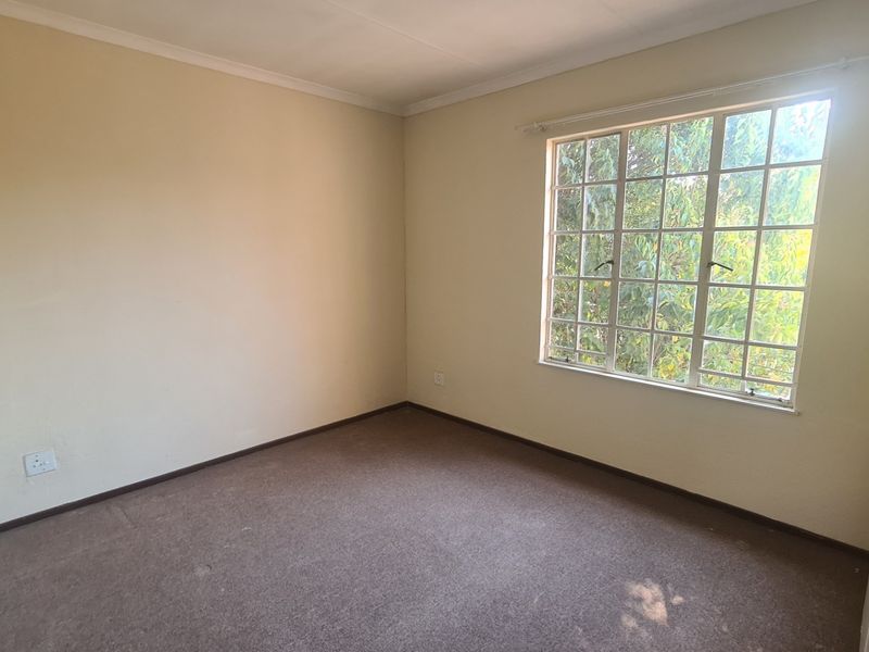 A vacant bedroom featuring neutral walls, carpeted flooring, a built-in cupboard, and a window with security bars overlooking greenery.
