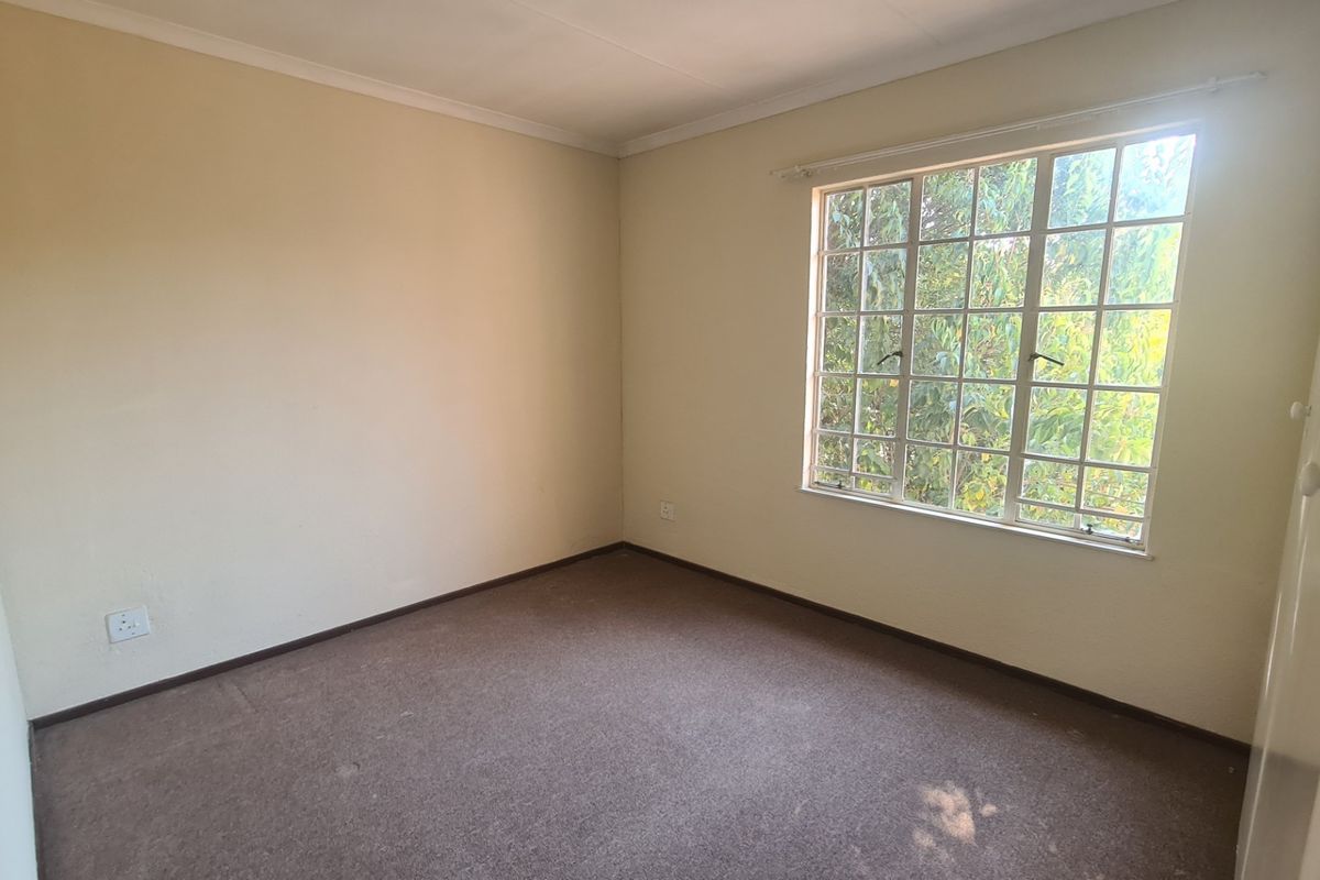 A vacant bedroom featuring neutral walls, carpeted flooring, a built-in cupboard, and a window with security bars overlooking greenery.