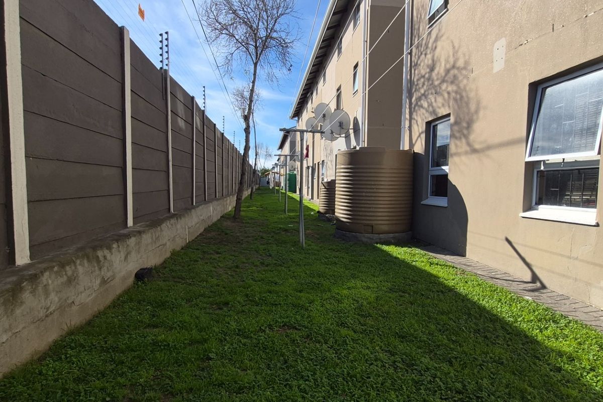 Rear exterior view of the apartment building featuring a grassed area, boundary wall with electric fencing, and installed water storage tanks.