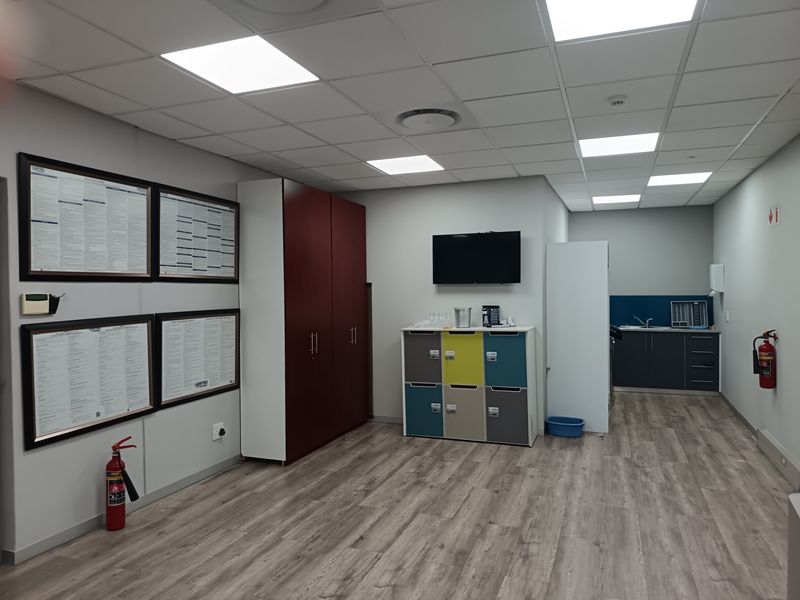 A modern office space featuring a kitchenette, built-in storage cabinetry, suspended ceiling with LED lighting, and durable laminate flooring.