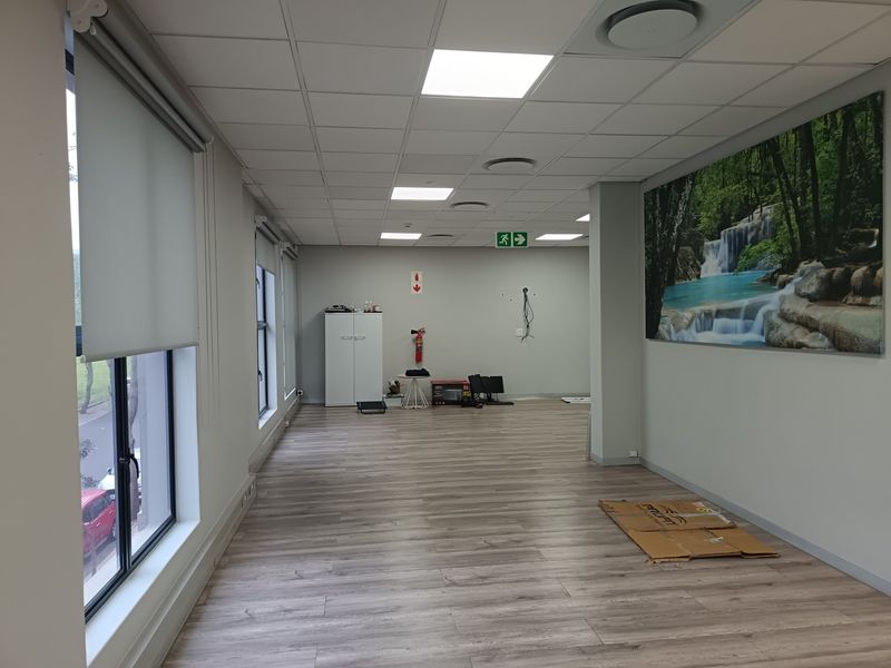 A spacious, vacant office area featuring modern laminate flooring, suspended ceilings with recessed lighting, and large windows with roller blinds.