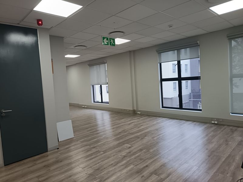 A bright, open-plan office space featuring modern laminate flooring, suspended ceilings with recessed lighting, and large windows with roller blinds.