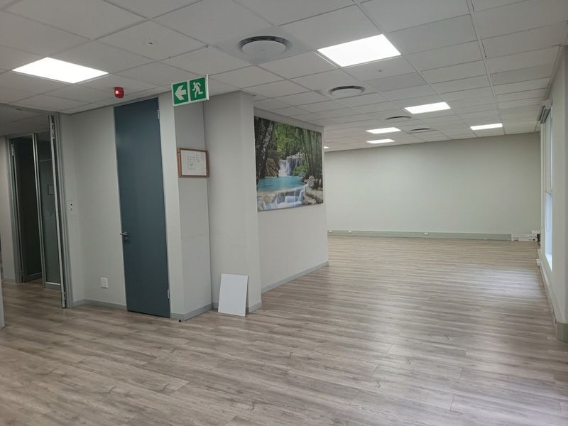 A spacious, vacant open-plan office area featuring modern laminate flooring, a suspended ceiling with recessed lighting, and integrated air conditioning.