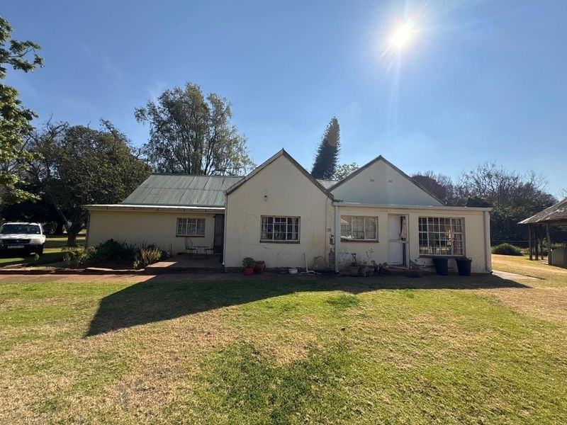 Rear view of a single-story farmhouse featuring a corrugated metal roof, plastered masonry walls, and a paved patio area set within a large garden.