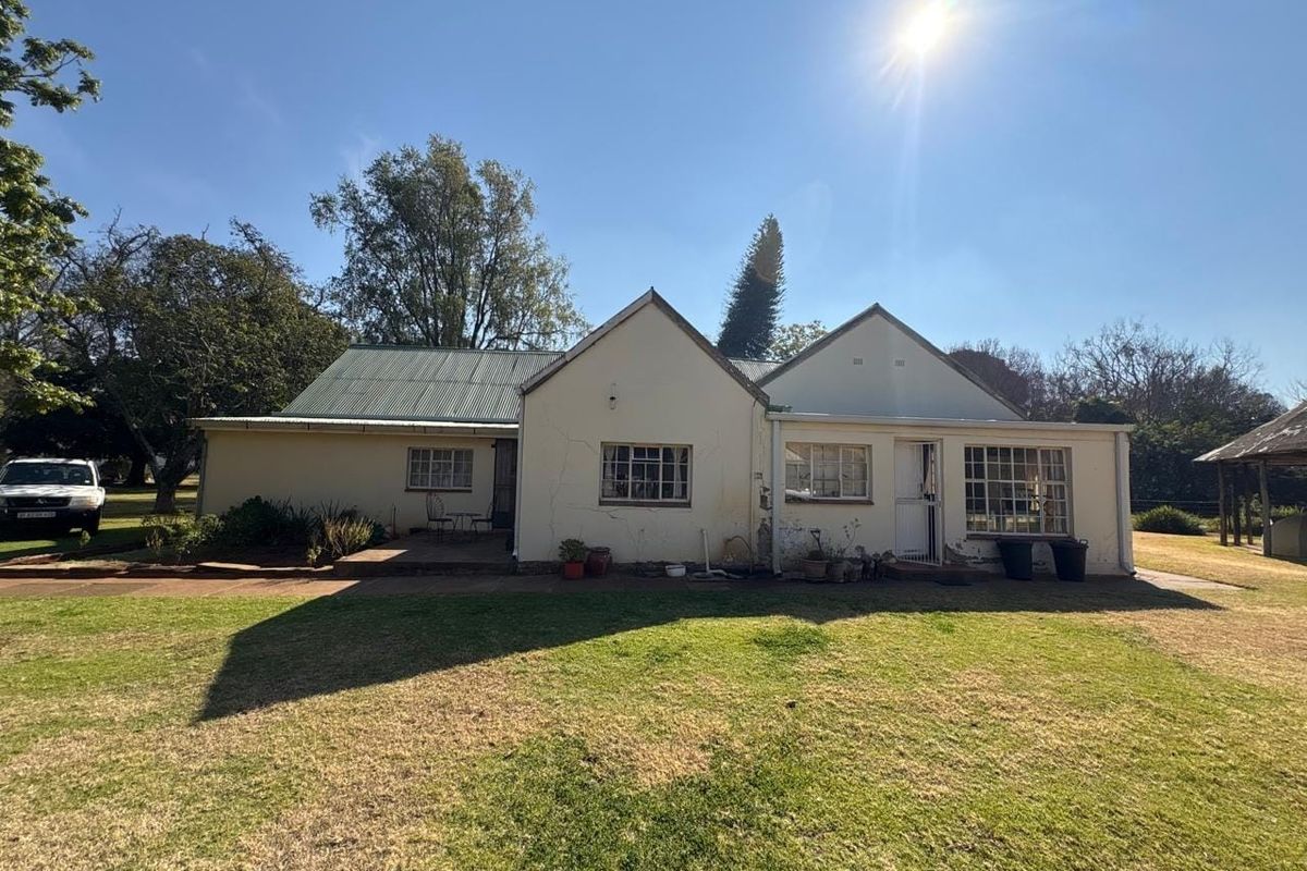 Rear view of a single-story farmhouse featuring a corrugated metal roof, plastered masonry walls, and a paved patio area set within a large garden.