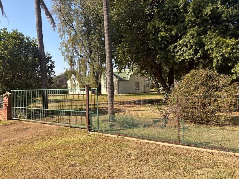 View of the farm property entrance featuring a metal security gate, wire fencing, and a glimpse of the main dwelling with a pitched roof.