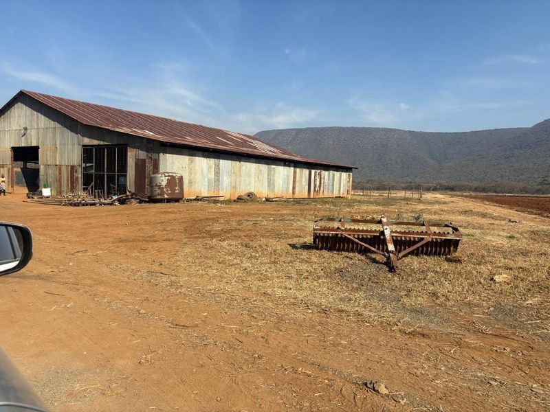 A large corrugated metal barn structure situated on a rural farm property with mountain views in the background.