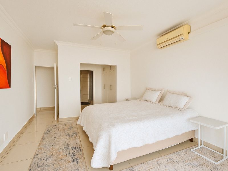 A bright bedroom featuring tiled flooring, built-in cupboards, a ceiling fan, and a wall-mounted air conditioning unit.