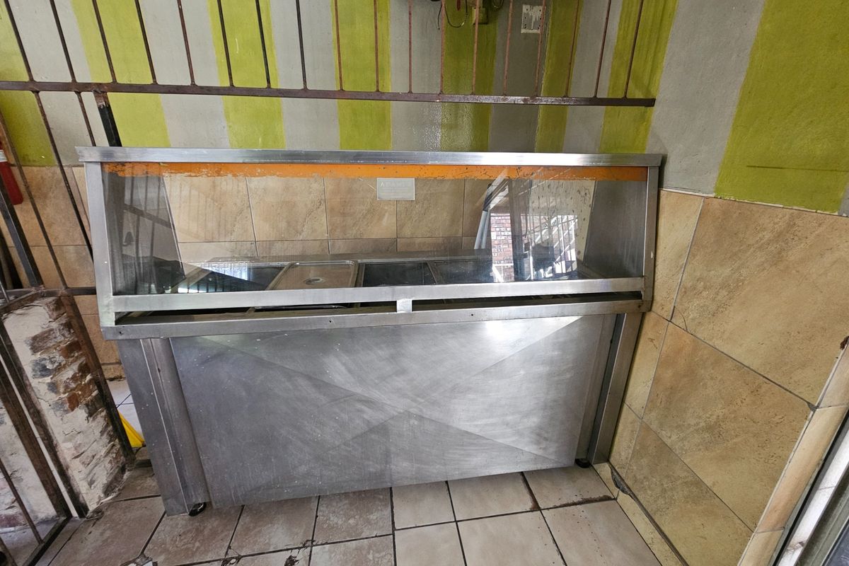 A utilitarian kitchen area featuring a stainless steel food display counter, tiled wall finishes, and tiled flooring, requiring cosmetic updates.
