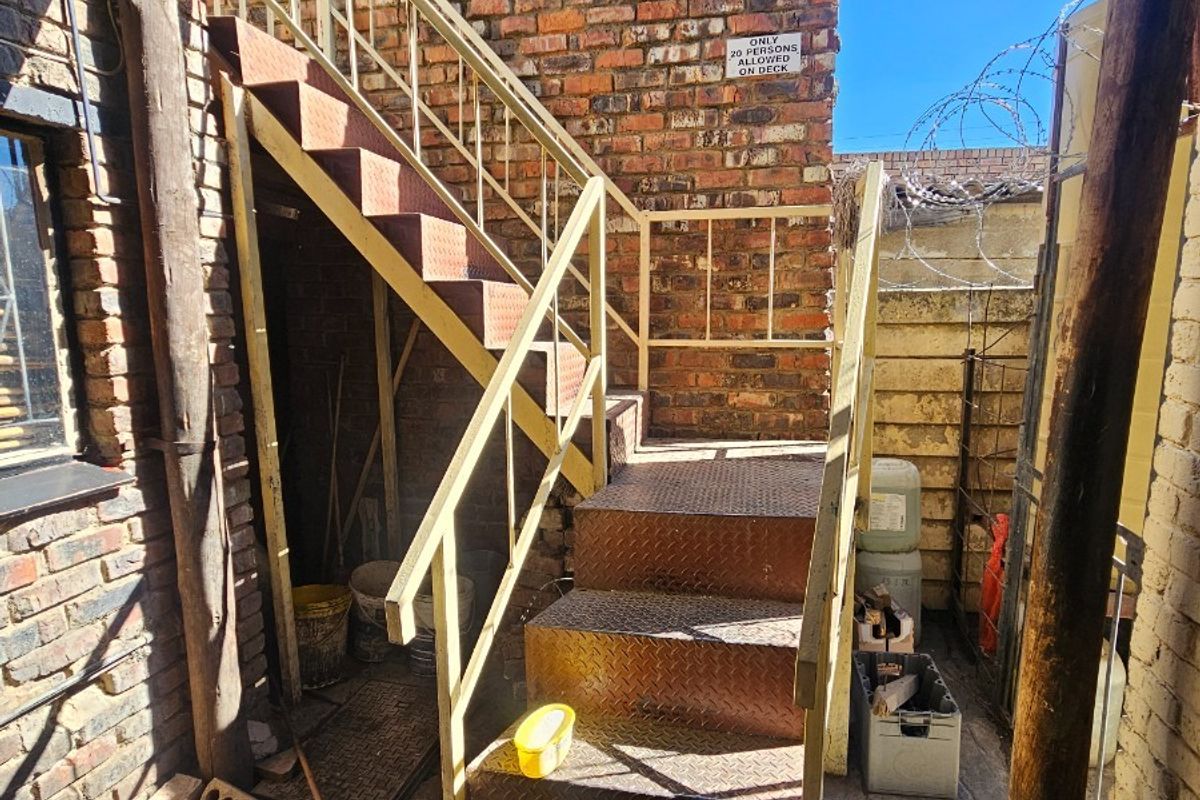 An exterior rear area featuring a metal staircase against a brick wall, with stone paving and security fencing.