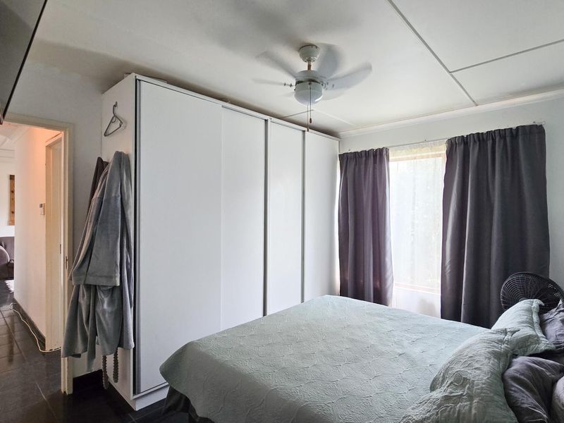A bedroom featuring a large built-in wardrobe, ceiling fan, and tiled flooring, offering a functional and neutral living space.