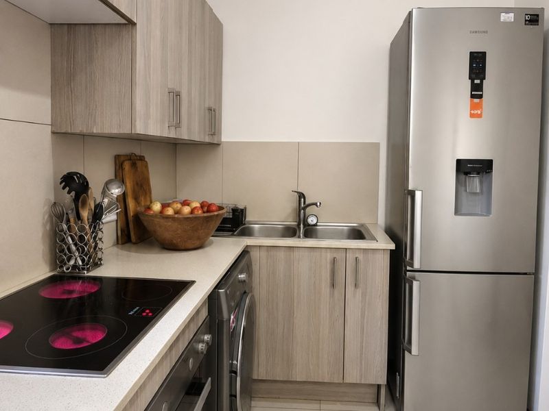 A modern, compact kitchen featuring light wood-grain cabinetry, stone countertops, integrated electric hob, built-in oven, and a double bowl sink with tiled flooring.