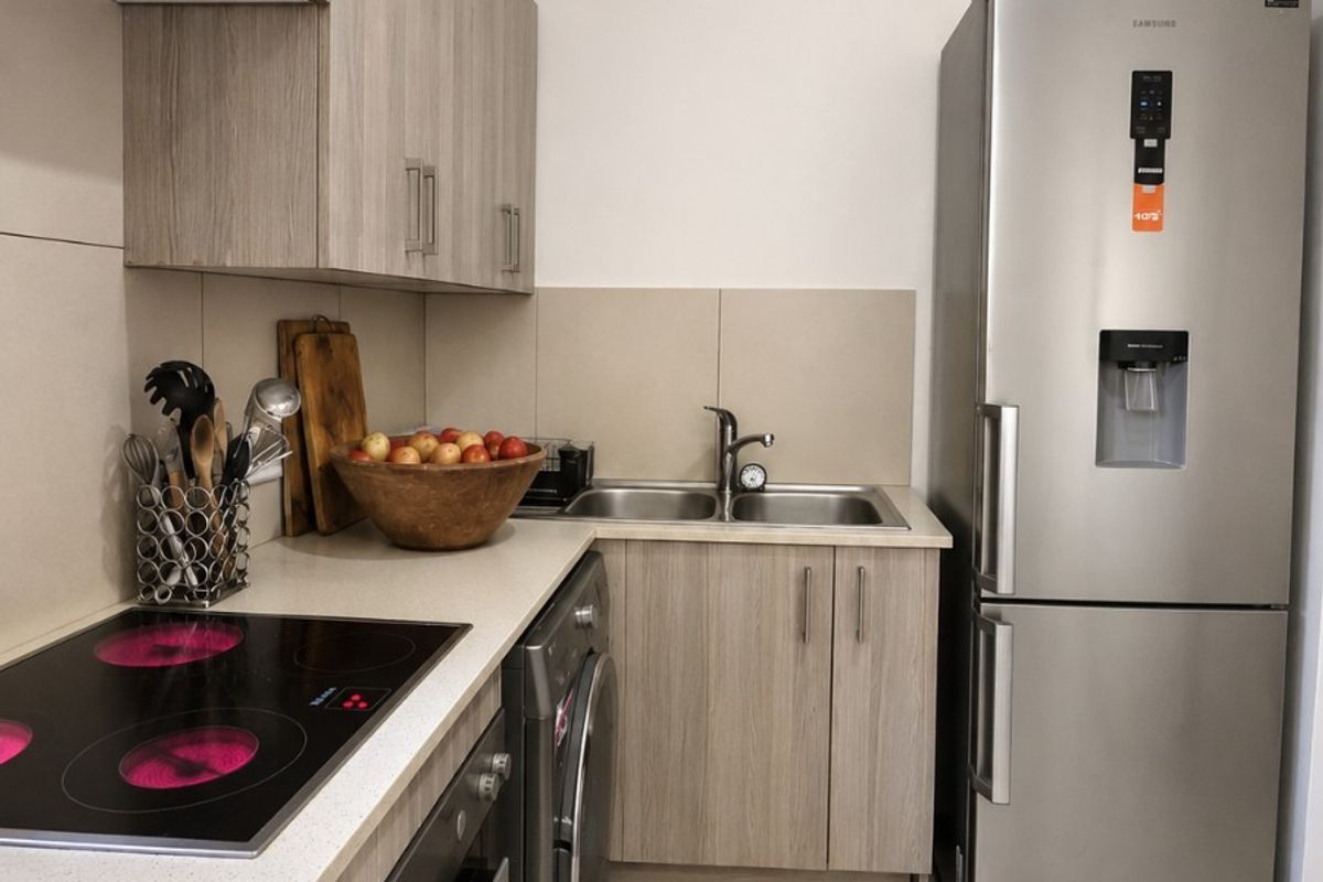 A modern, compact kitchen featuring light wood-grain cabinetry, stone countertops, integrated electric hob, built-in oven, and a double bowl sink with tiled flooring.