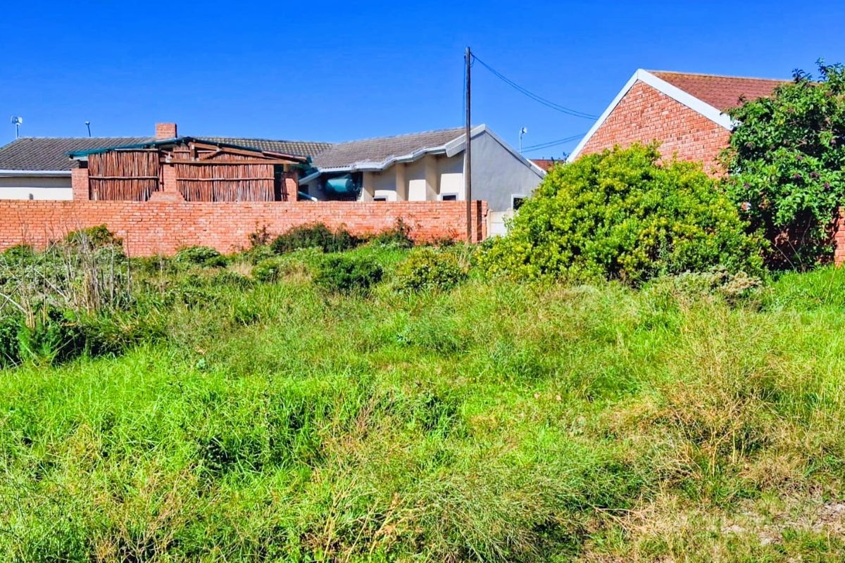 A view of a vacant residential plot featuring overgrown grass, a brick boundary wall, and adjacent residential structures.
