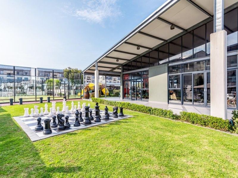 Modern clubhouse exterior featuring a covered patio, expansive glass facade, and a landscaped garden area with a large outdoor chess set.